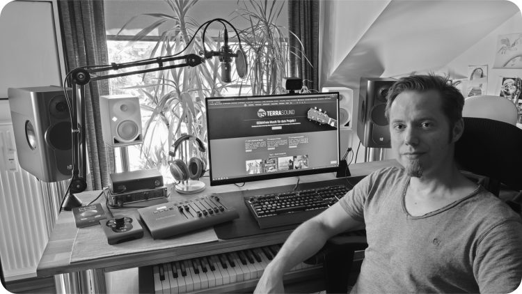 TerraSound - Tonstudio: Musikproduktionsstätte von Dag Reinbott. Das Bild zeigt den Arbeitsplatz mit Aktiv-Monitoren von Kii Audio und Neumann, ein Mikrofon am Teleskop-Arm, Audio-interface von RME, Midi-Mischpult, Tastatur und Bildschirm, Master-Keyboard unter dem Tisch verbaut. Im Vordergrund rechts sitzt Dag Reinbott.